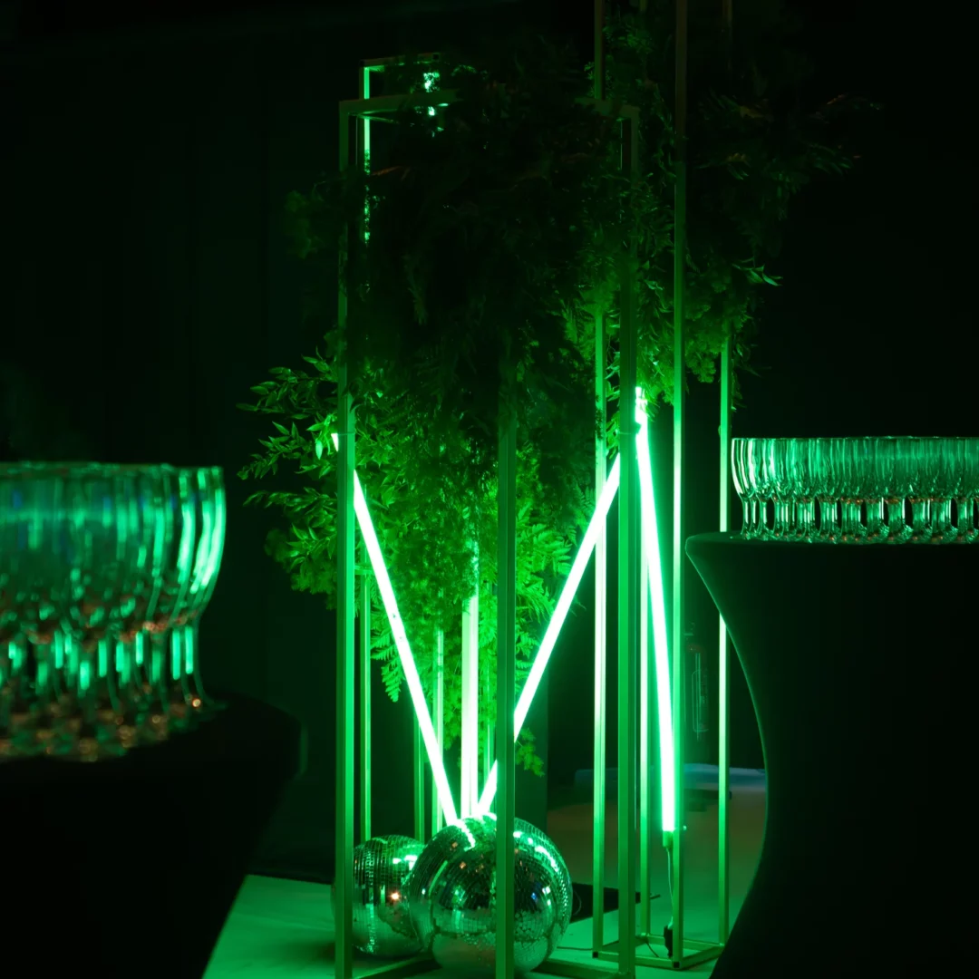 Nowoczesna scenografia eventowa z metalowymi słupami w złotym kolorze, zielonym neonowym światłem i roślinnością. Kule dyskotekowe u podstawy i stół z kieliszkami podkreślają elegancki klimat bankietowy. Idealna dekoracja na wesela, eventy i imprezy tematyczne. Dekoracje dostępne w wypożyczalni rent4events.pl oraz rent4weddings.pl, Wrocław - rent4events.pl wypożycz wrażenia, rent4weddings.pl zachowaj wspomnienia. Fot. @karolinakiec_fotografia.