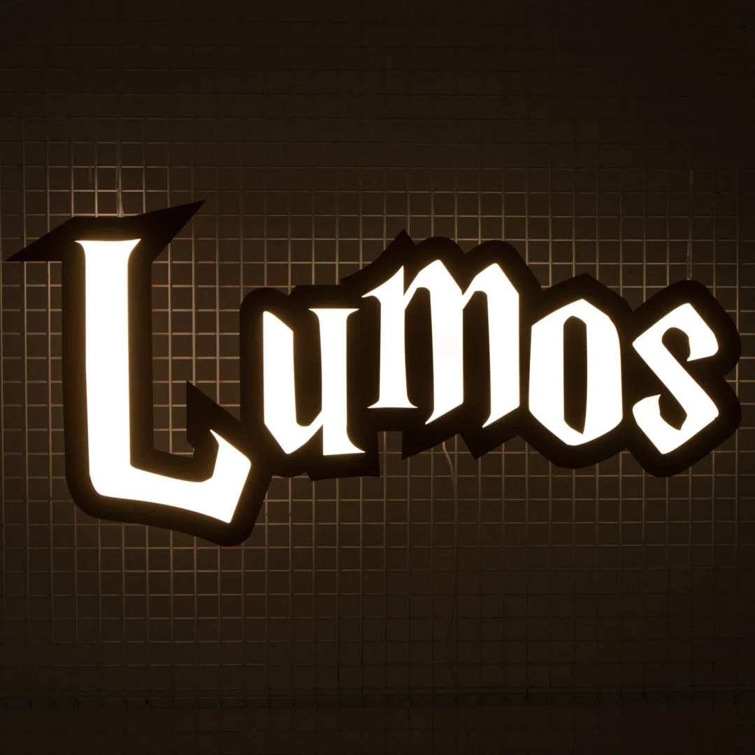 Neon Lumos - podświetlony napis „Lumos” wykonany gotycką czcionką na ciemnym tle. Jasne, białe litery emanują światłem, tworząc silny kontrast z ciemnym otoczeniem. Napis jest wyraźnie widoczny i może być częścią scenografii, dekoracji eventowej lub instalacji świetlnej o tematyce związanej z Harrym Potterem, fantasy, nawiązującej do magicznego świata. Idealne tło na event czy tematyczne wesele we Wrocławiu, szczególnie w klimacie fantasy. Scenografia inspirowana magią i tajemnicą, doskonała do stworzenia unikalnej atmosfery - rent4events.pl wypożycz wrażenia, rent4weddings.pl zachowaj wspomnienia.