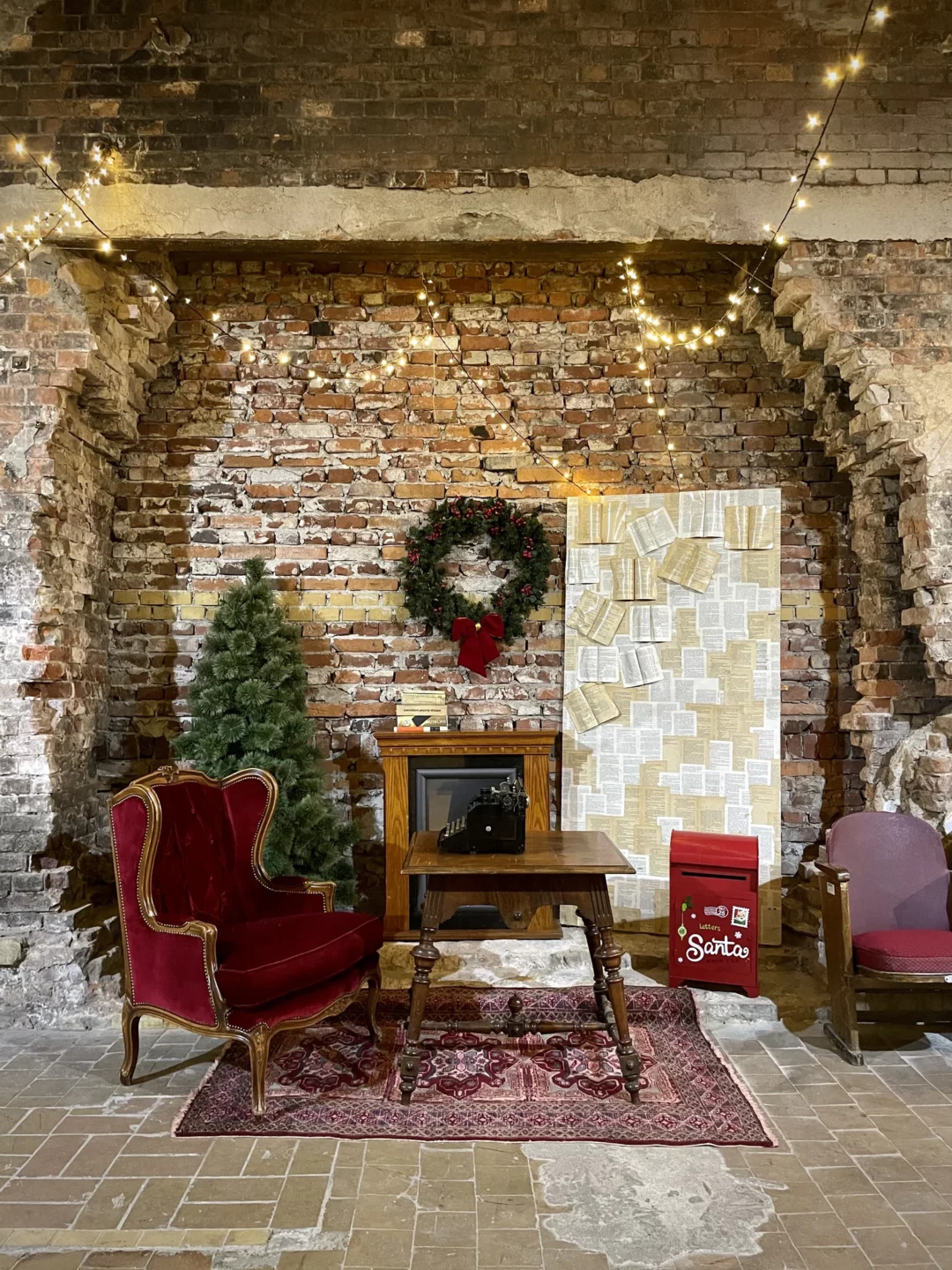 Stylizowana scenografia świąteczna z ceglaną ścianą, ozdobiona świątecznym wieńcem, choinką oraz dekoracjami retro. Bordowy tron z tapicerką vintage, drewniany stół z antyczną maszyną do pisania oraz tablica ze stron książek tworzą niepowtarzalny klimat. Girlandy świetlne dodają ciepłego nastroju. Dekoracje takie jak czerwona skrzynka na listy "Letters to Santa" i orientalny dywan idealnie komponują się w całość. Wypożyczalnia dekoracji świątecznych na eventy we Wrocławiu – rent4events.pl i rent4weddings.pl - rent4events.pl wypożycz wrażenia, rent4weddings.pl zachowaj wspomnienia. Fot. Martyna.