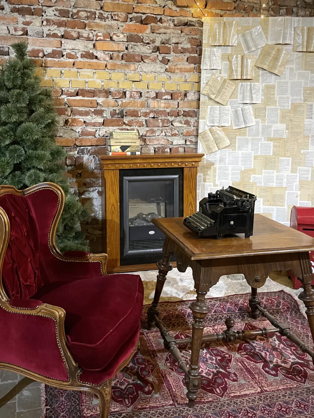 Stylowa świąteczna aranżacja wnętrza z ceglanym tłem i retro dekoracjami. Centralnym punktem jest drewniany kominek z antyczną maszyną do pisania na rustykalnym stole. Obok znajduje się czerwone, tapicerowane krzesło - Tron Bordowy - o klasycznym wyglądzie oraz zielona choinka. W tle widoczna tablica z przyklejonymi stronami książek, tworząca unikalną scenografię. Całość dopełnia orientalny dywan w odcieniach czerwieni. Wypożyczalnia dekoracji na ślub, wesele czy event we Wrocławiu – rent4events.pl i rent4weddings.pl - rent4events.pl wypożycz wrażenia, rent4weddings.pl zachowaj wspomnienia. Fot. Martyna.