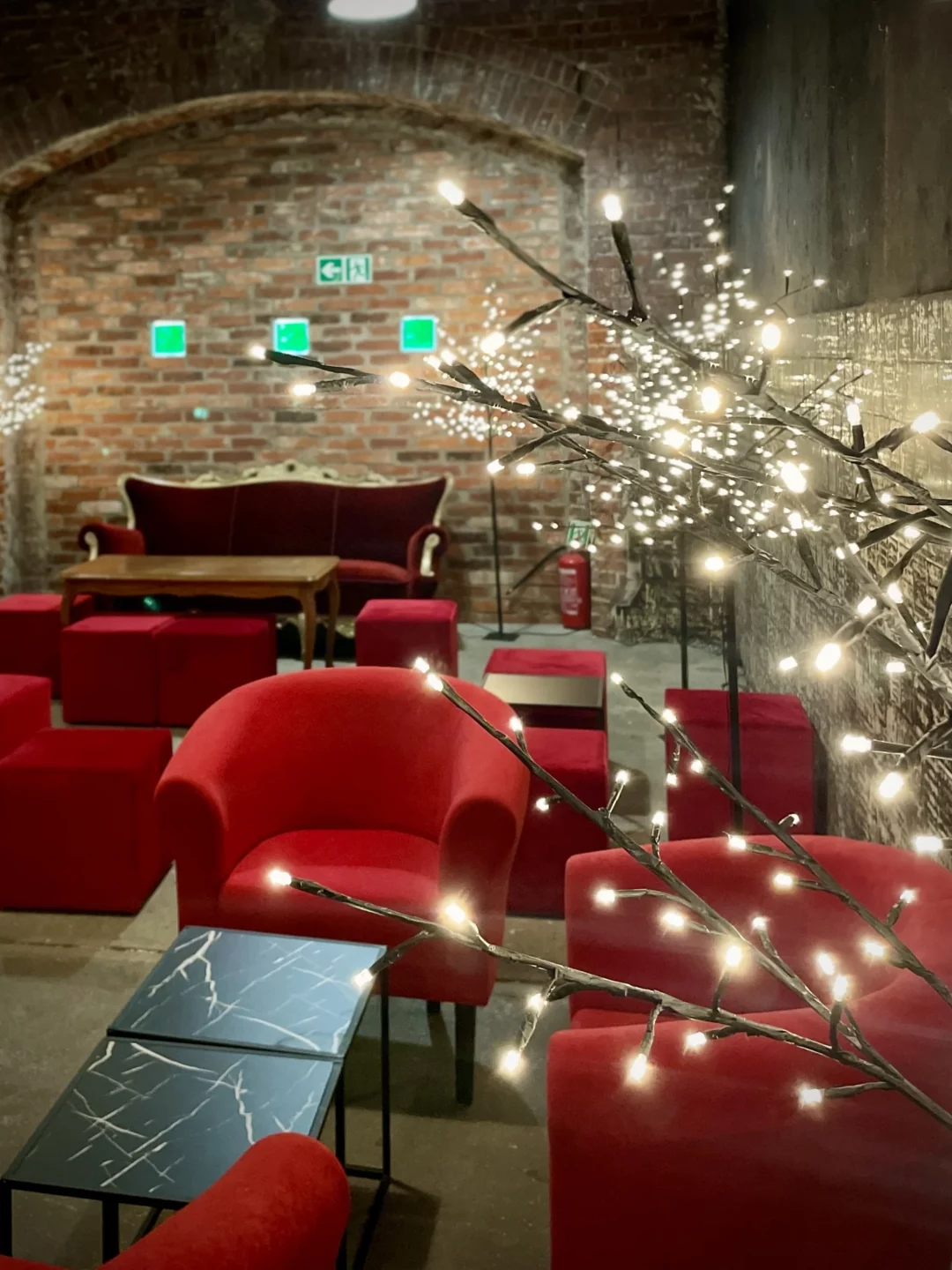 Czerwony chillout i Drzewka Led. Industrialna przestrzeń eventowa z ceglaną ścianą w tle. Elegancka czerwona sofa i fotele o aksamitnej tapicerce kontrastują z betonowym podłożem. W otoczeniu znajdują się sześcienne, czerwone pufy oraz nowoczesne, czarne stoliki. Dekoracyjne drzewka LED z delikatnym oświetleniem tworzą magiczną atmosferę. Industrialne akcenty, jak oświetlenie sufitowe i surowa, ceglana struktura, nadają wnętrzu unikalny charakter. Wypożyczalnia dekoracji i scenografii na event, ślub oraz wesele we Wrocławiu - rent4events.pl wypożycz wrażenia, rent4weddings.pl zachowaj wspomnienia. Fot. Matyna.