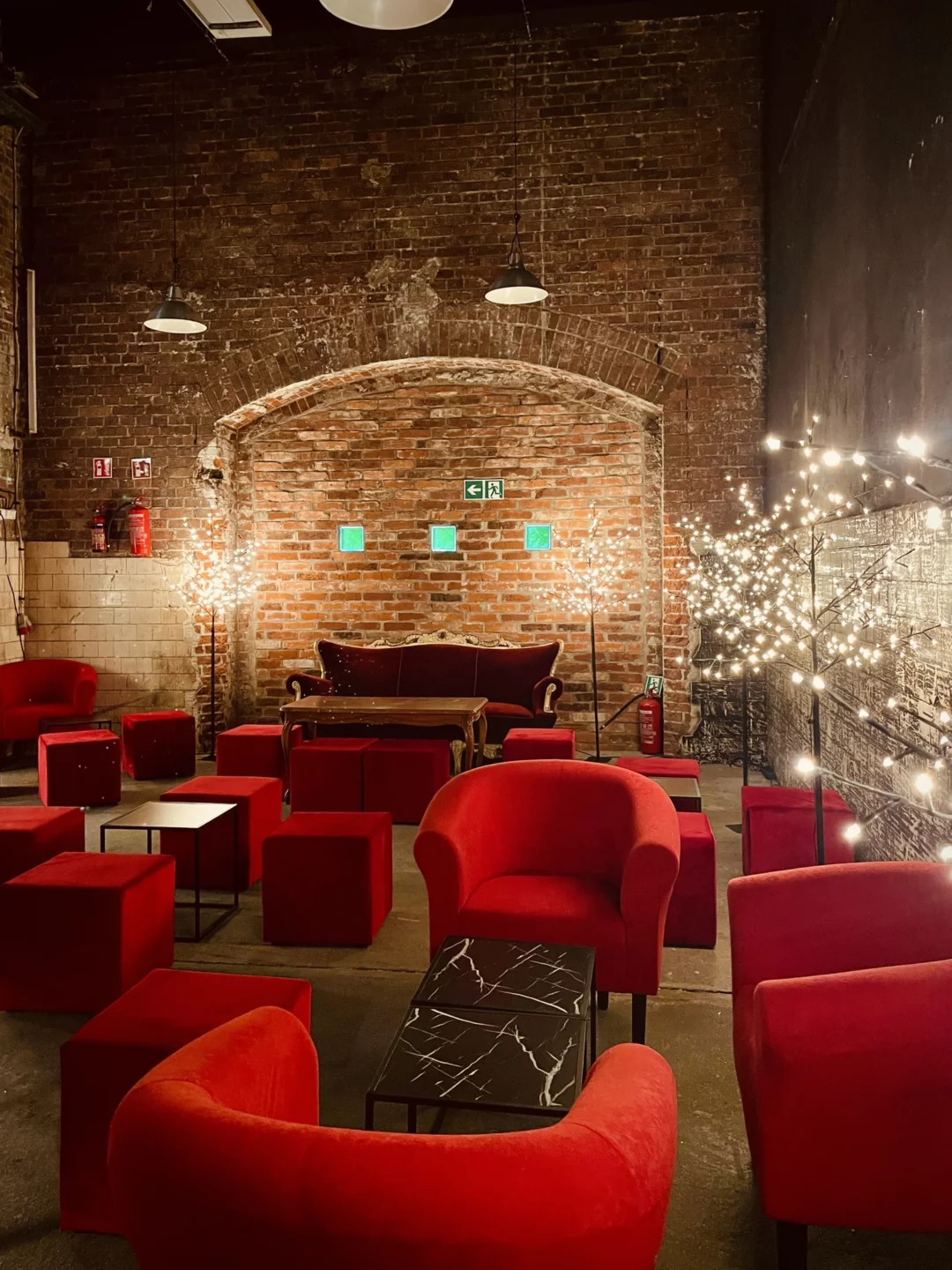 Czerwony chillout i Drzewka Led. Industrialna aranżacja eventowa z ceglaną ścianą w tle. Główne elementy to elegancka czerwona sofa, okrągłe fotele z aksamitną tapicerką oraz liczne sześcienne pufy w głębokim czerwonym odcieniu. Dekoracyjne drzewka LED z delikatnym światłem dodają klimatu, kontrastując z surowością loftowej przestrzeni. Czarny stolik z marmurowym wzorem dopełnia elegancję scenografii. Wypożyczalnia dekoracji na ślub, wesele i eventy we Wrocławiu - rent4events.pl wypożycz wrażenia, rent4weddings.pl zachowaj wspomnienia. Fot. Martyna.