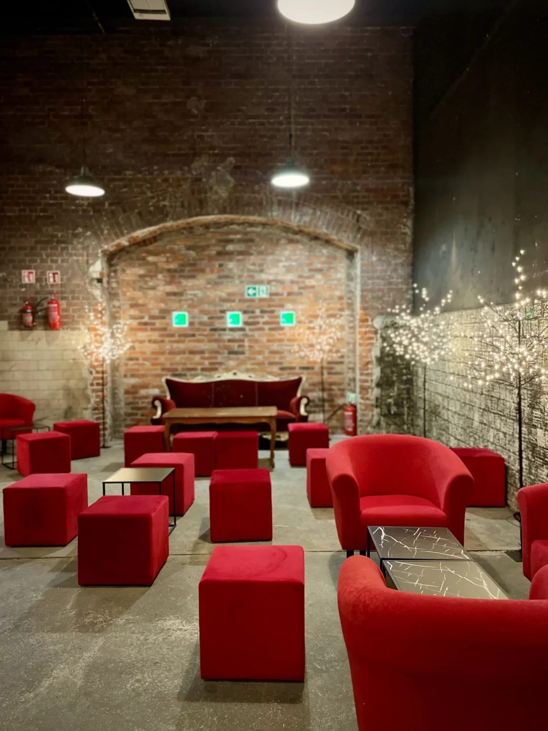 Czerwony chillout i Drzewka Led. Industrialna przestrzeń eventowa z ceglaną ścianą w tle. W centrum znajduje się elegancka czerwona sofa, otoczona licznymi czerwonymi sześciennymi pufami i okrągłymi fotelami o aksamitnej tapicerce. Dekoracyjne drzewka LED z delikatnym, białym oświetleniem podkreślają klimat wnętrza. Czarny, nowoczesny stolik z marmurowym wzorem dodaje elegancji przestrzeni. Surowy betonowy podkład oraz oświetlenie loftowe tworzą wyjątkową industrialną scenografię. Wypożyczalnia dekoracji na eventy, wesela i śluby we Wrocławiu – rent4events.pl wypożycz wrażenia, rent4weddings.pl zachowaj wspomnienia. Fot. Martyna.