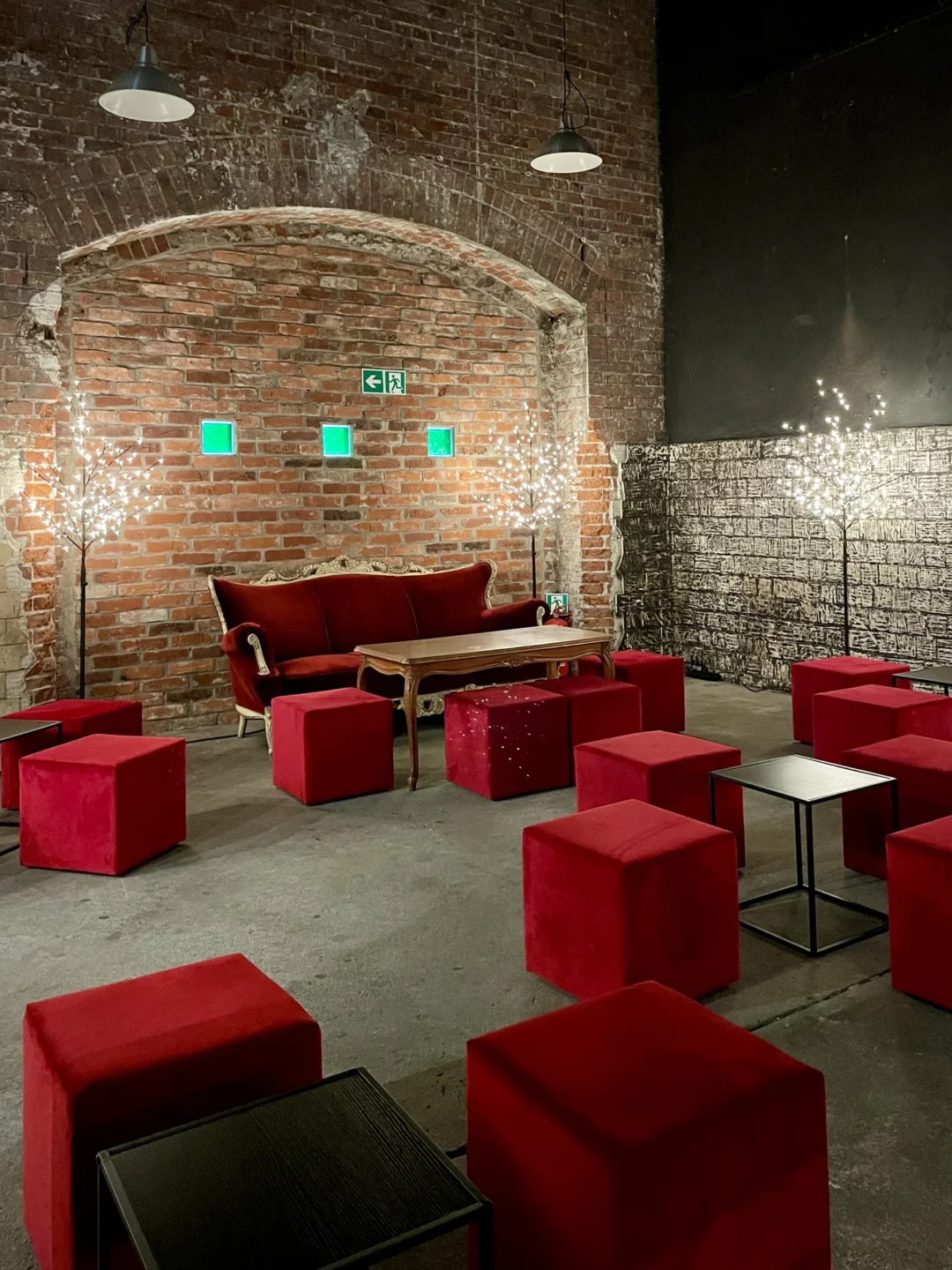 Czerwony chillout. Stylowa industrialna scenografia eventowa z ceglaną ścianą w tle. Centralnie umieszczona elegancka, czerwona sofa w otoczeniu licznych tapicerowanych kostek siedziskowych w kolorze czerwonym. Całość dopełniają drewniany stolik retro oraz dekoracyjne podświetlane drzewka LED, dodające przytulnego klimatu. Oświetlenie sufitowe w stylu loftowym oraz surowe, betonowe podłoże podkreślają industrialny charakter aranżacji. Wypożyczalnia dekoracji i scenografii eventowej na ślub, wesele oraz eventy we Wrocławiu – rent4events.pl i rent4weddings.pl - rent4events.pl wypożycz wrażenia, rent4weddings.pl zachowaj wspomnienia. Fot. Martyna.