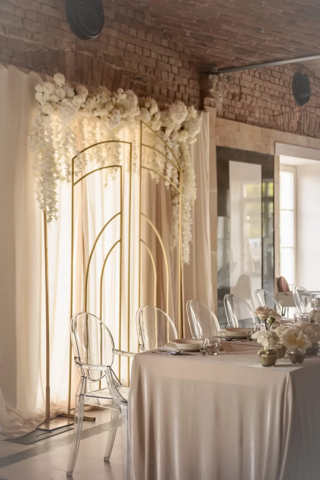 Stylowa aranżacja wnętrza na wesele lub event w stylu glamour we Wrocławiu. Złoty stelaż z geometrycznych łuków, ozdobiony białymi różami, hortensjami i kaskadami wisterii, wkomponowany w tło z jasnych, beżowych zasłon. Stół z eleganckim welurowym obrusem w szampańskim odcieniu, udekorowany kwiatami w złotych wazonach, dopełnia przezroczystymi krzesłami nowoczesny i minimalistyczny wystrój. Ceglana ściana nadaje przestrzeni industrialny charakter, tworząc unikalną scenografię. Idealna dekoracja ślubna lub eventowa od rent4events.pl - rent4events.pl – wypożycz wrażenia, rent4weddings.pl – zachowaj wspomnienia.