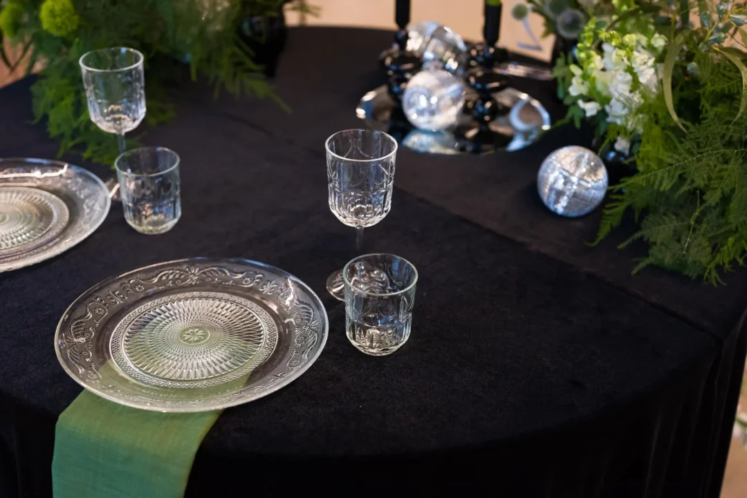 Ekskluzywna aranżacja stołu eventowego autorstwa wypożyczalni RENT4EVENTS / RENT4WEDDING z Wrocławia. Na czarnym, aksamitnym obrusie znajdują się kryształowe podtalerze i kieliszki o szlifie vintage, tworzące eleganckie nakrycie. Dekorację uzupełnia zielona serweta, subtelna florystyka z białymi kwiatami i srebrne kule dyskotekowe umieszczone przy świecznikach na lustrzanej tacy. Idealna scenografia na nowoczesny ślub, wesele czy stylowy event w styylu disco. Całość dostępna do wypożyczenia na stronie rent4events.pl oraz w ofercie ślubnej marki rent4weddings.pl - rent4events.pl wypożycz wrażenia, rent4wedding.pl zachowaj wspomnienia. Fot. Maciej Wójcik Fotograf.
