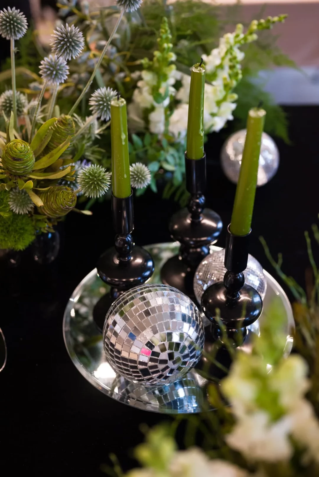 Dekoracja stołu w stylu glamour stworzona przez wypożyczalnię RENT4EVENTS / RENT4WEDDING, łącząca zielone świece w czarnych, eleganckich świecznikach z błyszczącymi kulami dyskotekowymi umieszczonymi na lustrzanej tacy. Aranżacja wzbogacona bujnymi kompozycjami roślinnymi z mikołajkami, lwiemi paszczami i sukulentami w intensywnych zielono-białych barwach. Stylowa, nowoczesna aranżacja stołu doskonale sprawdzi się na wesele, ślub lub event we Wrocławiu. Zestaw dostępny w ofercie wypożyczalni dekoracji rent4events.pl oraz rent4weddings.pl – idealny wybór dla osób poszukujących nietuzinkowej oprawy wizualnej - rent4events.pl wypożycz wrażenia, rent4wedding.pl zachowaj wspomnienia. Fot. Maciej Wójcik Fotograf.