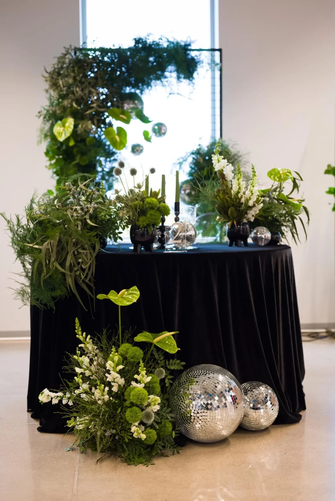 Elegancka scenografia eventowa zaaranżowana przez wypożyczalnię dekoracji RENT4EVENTS / RENT4WEDDING prezentuje okrągły stół udekorowany w nowoczesnym stylu z czarnym aksamitnym obrusem z weluru, z zielonymi kompozycjami roślinnymi oraz srebrnymi kulami dyskotekowymi w stylu glamour. Na stole znajdują się zielono-białe bukiety z anturium, lwimi paszczami i mchami, umieszczone w czarnych wazonach. Tło stanowi przestrzenna konstrukcja z bujną zielenią – idealna na ślub, wesele lub wyjątkowy event we Wrocławiu. Dekoracje tworzą spójną, designerską całość, nadając przestrzeni prestiżowy charakter. Całość dostępna w ofercie marek rent4events.pl oraz rent4weddings.pl - rent4events.pl wypożycz wrażenia, rent4wedding.pl zachowaj wspomnienia. Fot. Maciej Wójcik Fotograf.