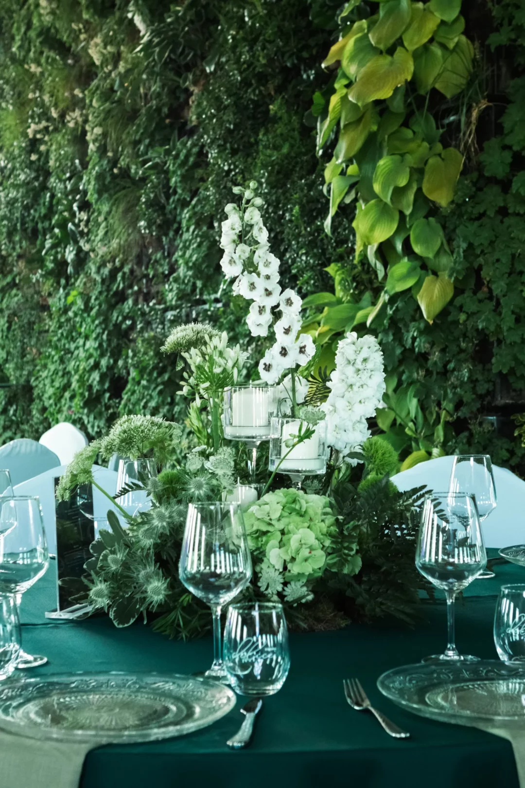 Elegancka dekoracja stołu z bujnymi zielonymi roślinami i białymi kwiatami, które nadają naturalny i świeży wygląd. Na stole znajdują się przezroczyste, szklane świeczniki na nóżce z białymi swiecami, kryształowe podtalerze oraz szklane kieliszki i szklanki, które dodają lekkości aranżacji, a świece w szklanych świecznikach wprowadzają subtelny, elegancki akcent. Zielone tło roślinne w tle tworzy harmonijną scenografię, idealną na wesele lub event we Wrocławiu. Aranżacja dostępna w wypożyczalni rent4events.pl oraz rent4weddings.pl. rent4events.pl wypożycz wrażenia, rent4wedding.pl zachowaj wspomnienia. Fot. @karolinakiec_fotografia.