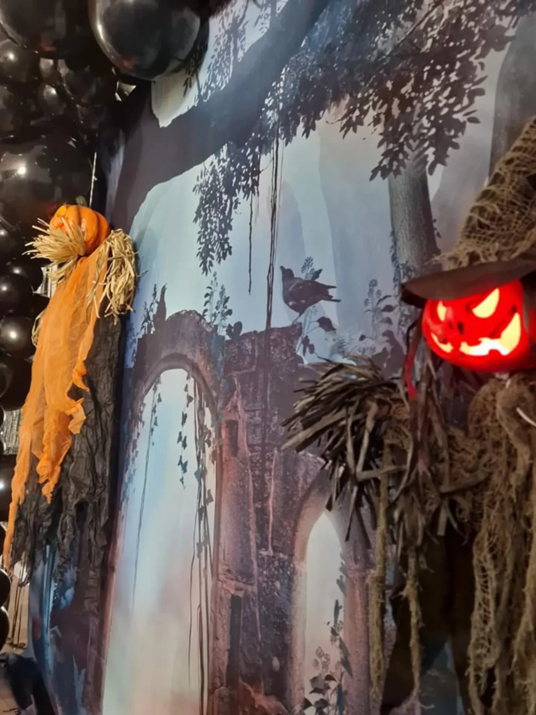 Dekoracja halloweenowa z tłem fotograficznym przedstawiającym mroczną scenerię lasu z ruinami łuku porośniętego bluszczem. Na pierwszym planie dynia z pomarańczową chustą oraz lampion w kształcie świecącej, upiornej głowy wyciętej w dyni. W tle czarne balony podkreślające tajemniczy klimat. Idealna scenografia na tematyczne eventy we Wrocławiu - rent4events.pl wypożycz wrażenia, rent4weddings.pl zachowaj wspomnienia.