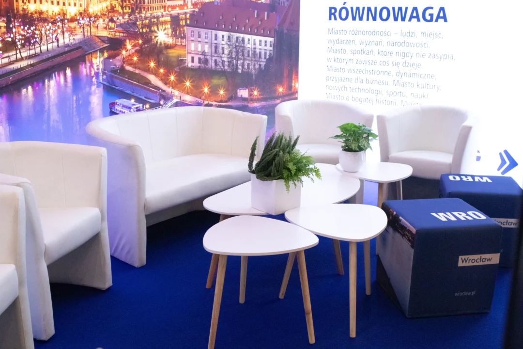 Elegancka aranżacja strefy wypoczynkowej/kawowej z białymi fotelami i sofą, uzupełniona o nowoczesne stoliki na drewnianych nogach oraz zielone rośliny w minimalistycznych doniczkach. Sceneria uzupełniona jest podświetlonym tłem z widokiem na panoramę Wrocławia. Niebieskie pufy z nadrukiem „WRO” dodają nowoczesnego charakteru i promują miasto. Doskonała scenografia na eventy biznesowe i promocyjne. Stylowe wyposażenie dostępne w ofercie wypożyczalni Rent4Events - rent4events.pl wypożycz wrażenia, rent4weddings.pl zachowaj wspomnienia. Fot. @karolinakiec_fotografia.