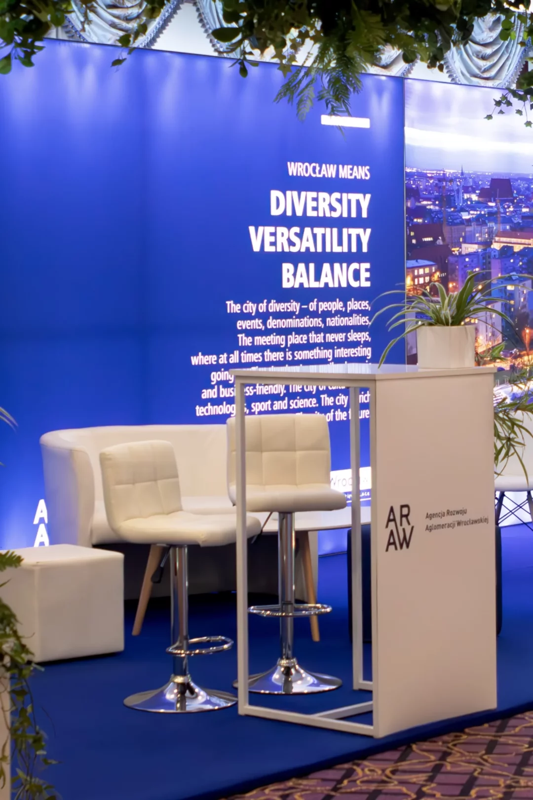 Nowoczesna przestrzeń eventowa zaprojektowana w stylu biznesowym z dominującym niebieskim tłem i napisem „Wrocław Means Diversity, Versatility, Balance”. W centrum znajduje się elegancka, biała strefa powitalna z wysokim stolikiem i wygodnymi hokerami o chromowanych podstawach, podkreślających minimalistyczny design. Z tyłu widoczna biała sofa strefy wypoczynkowej. Scenografia idealna na wydarzenia biznesowe, konferencje i eventy promujące Wrocław. Dekoracje i wyposażenie dostępne w Rent4Events - rent4events.pl wypożycz wrażenia, rent4weddings.pl zachowaj wspomnienia. Fot. @karolinakiec_fotografia.