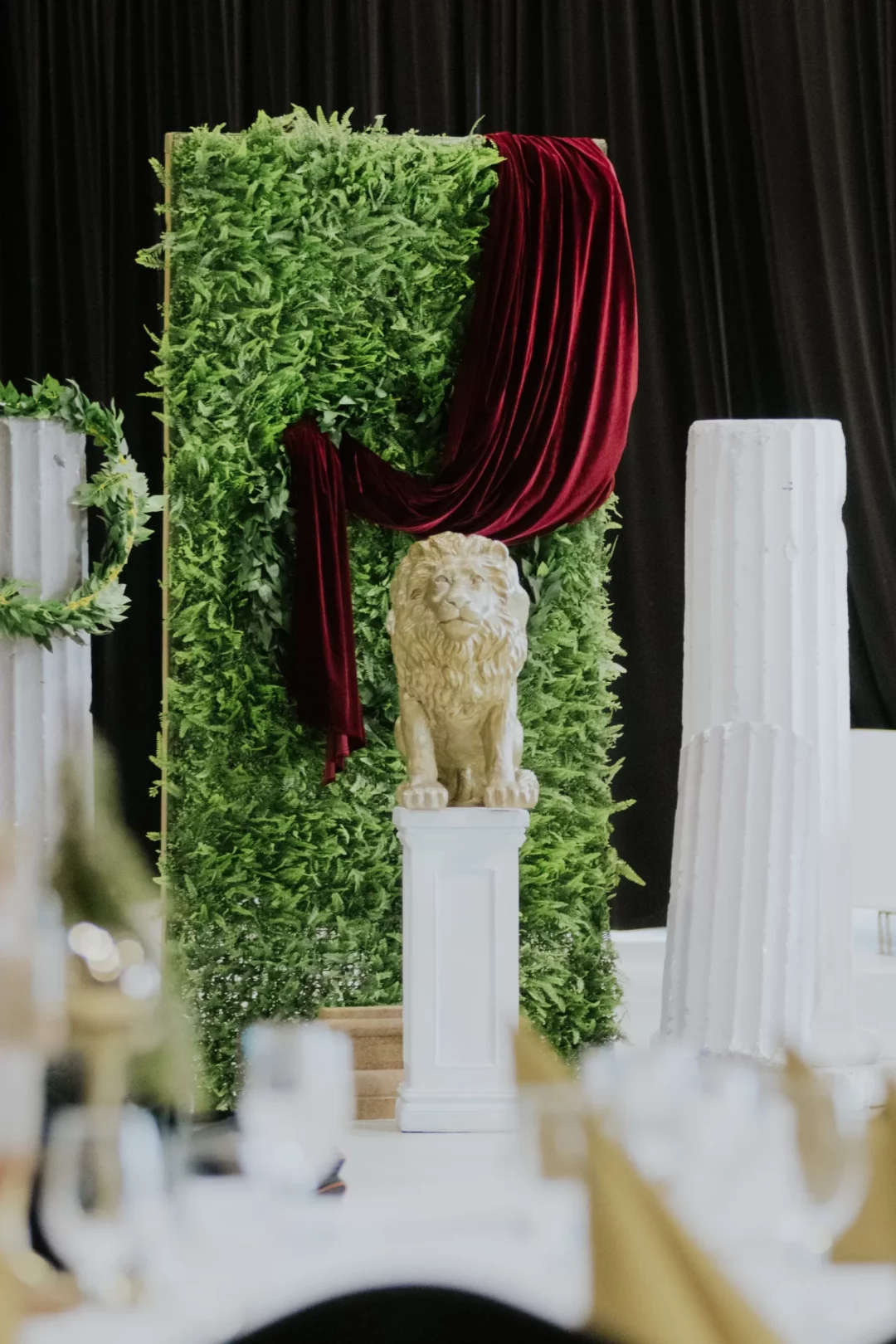 Scenografia z rzymskimi kolumnami, złotym lwem i ścianą pokrytą zielenią, doskonała na eventyw tematyce "Opowieści z Narnii" oraz zimowe uroczystości w baśniowym klimacie, we Wrocławiu i okolicach. Centralnym elementem jest figura złotego lwa na białym postumencie, ustawiona na tle zieleni z bordowym aksamitnym materiałem. Kolumny w stylu rzymskim dodają klasycznej elegancji i antycznego uroku, idealnie wpasowując się w scenografię glamour. Dekoracje oferowane są przez wypożyczalnię rent4events.pl oraz rent4weddings.pl - rent4events.pl wypożycz wrażenia, rent4wedding.pl zachowaj wspomnienia. Fot. @karolinakiec_fotografia.