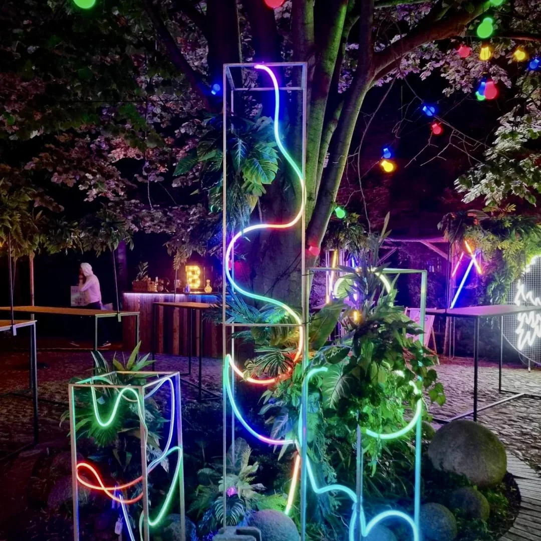 Słupek LED 250 cm, 150 cm i 100 cm. Neon Party. Efektowna dekoracja na event, tu w plenerze, przedstawiająca, zamontowane na trzech srebrnych słupkach różnej wysokości, neonowe struktury w kształcie abstrakcyjnych linii, otoczone bujną roślinnością i delikatnym światłem dekoracyjnym. W tle widać bar oraz ozdobne żarówki zawieszone na drzewach, które tworzą magiczną atmosferę idealną na ślub, wesele lub inny event. Neonowe kolory – różowy, zielony, niebieski i czerwony – wyróżniają się na tle wieczornego nieba i dodają scenografii wyjątkowego charakteru. Elementy dekoracyjne dostępne do wypożyczenia na stronie rent4events.pl i rent4weddings.pl - rent4events.pl wypożycz wrażenia, rent4weddings.pl zachowaj wspomnienia. Fot. Karol.