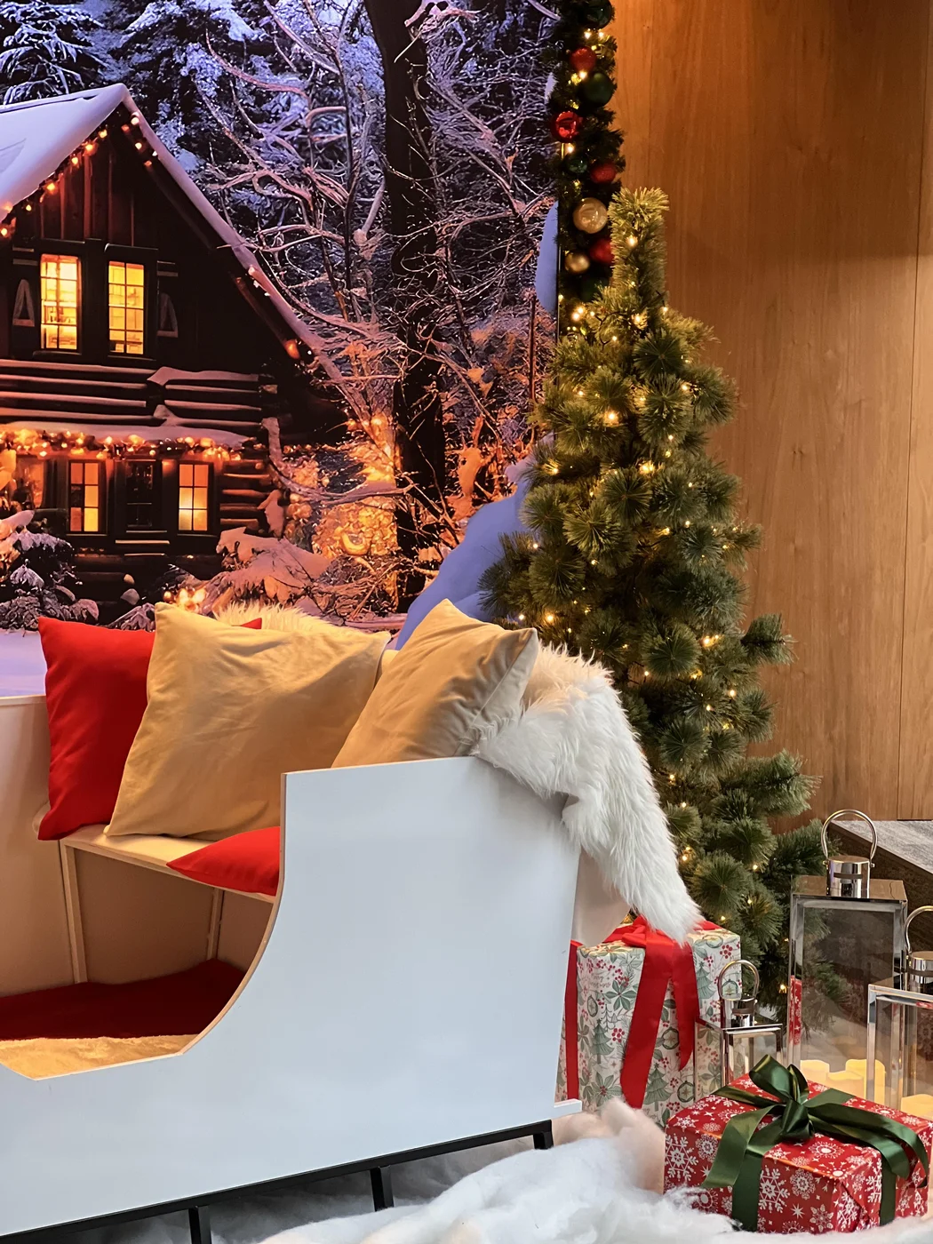 Świąteczna scenografia z białymi saniami dekoracyjnymi, czerwonymi i beżowymi poduszkami, futrzaną narzutą oraz stylizowanymi prezentami ustawionymi na sztucznym śniegu, uzupełniona choinką LED i metalowymi lampionami, na tle zdjęcia zimowego domku z bali w leśnej aranżacji – klimatyczne dekoracje idealne na firmowy event czy zimowy ślub. Profesjonalna wypożyczalnia z miasta Wrocław oferuje kompleksową realizację, montaż i transport scenografii dostępnej w ofercie rent4events.pl oraz rent4weddings.pl, tworząc efektowne strefy foto i wyjątkowe przestrzenie eventowe - rent4events.pl wypożycz wrażenia, rent4wedding.pl zachowaj wspomnienia.