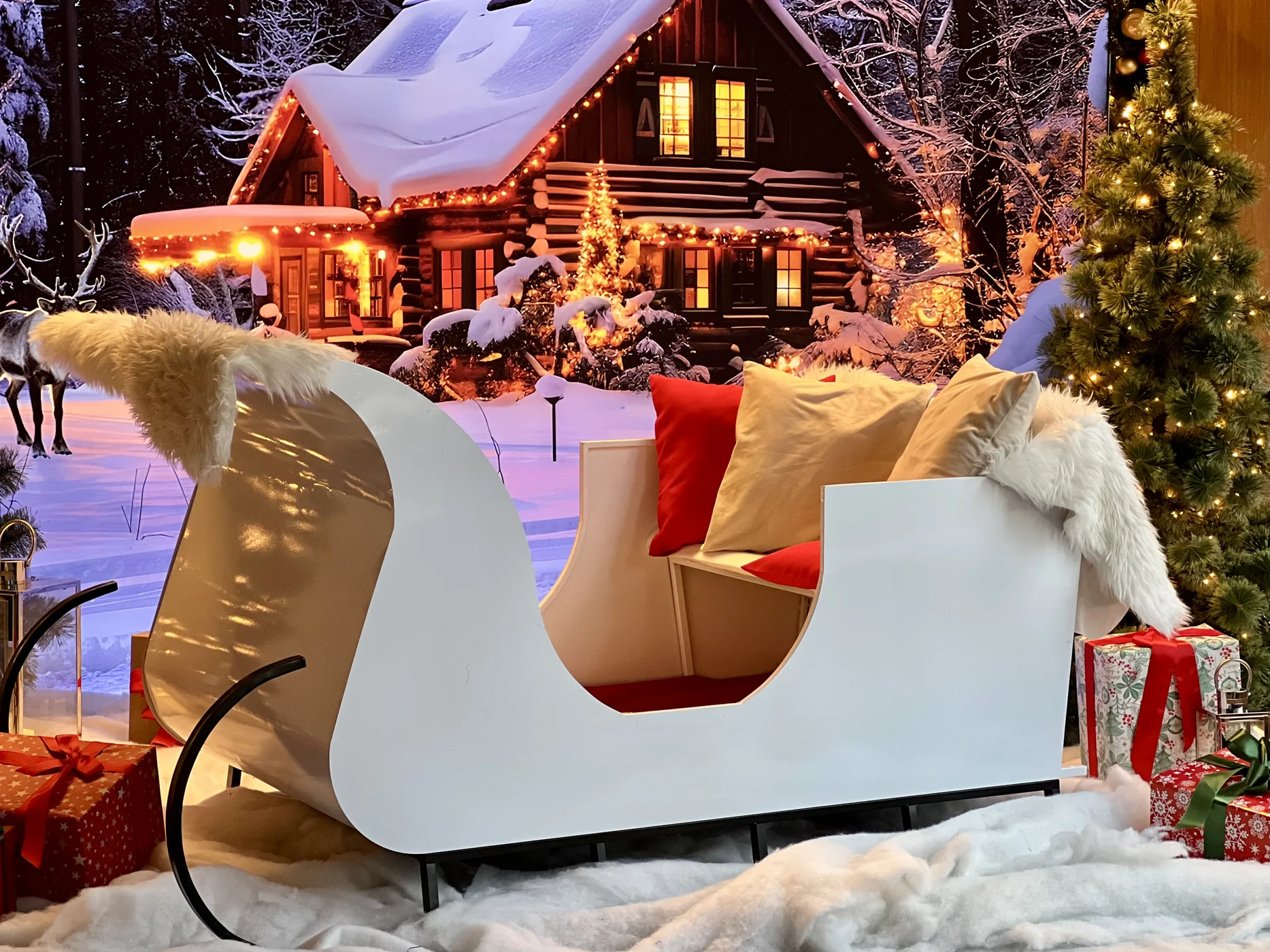 Zimowa scenografia świąteczna z białymi saniami dekoracyjnymi, czerwonymi i beżowymi poduszkami, futrzanymi narzutami oraz stylizowanymi prezentami ustawionymi na sztucznym śniegu, w tle tło fotograficzne z drewnianym domem z bali oświetlonym ciepłymi lampkami LED i choinką z iluminacją – klimatyczna aranżacja idealna jako dekoracje na event, firmową imprezę, jarmark, sesję zdjęciową, a także zimowy ślub i wesele. Profesjonalna wypożyczalnia scenografii z miasta Wrocław oferuje kompleksowe rozwiązania aranżacyjne od projektu po montaż. Ta bajkowa instalacja dostępna w ofercie rent4events.pl oraz rent4weddings.pl tworzy wyjątkowe tło fotograficzne i niezapomnianą atmosferę każdego wydarzenia - rent4events.pl wypożycz wrażenia, rent4wedding.pl zachowaj wspomnienia.