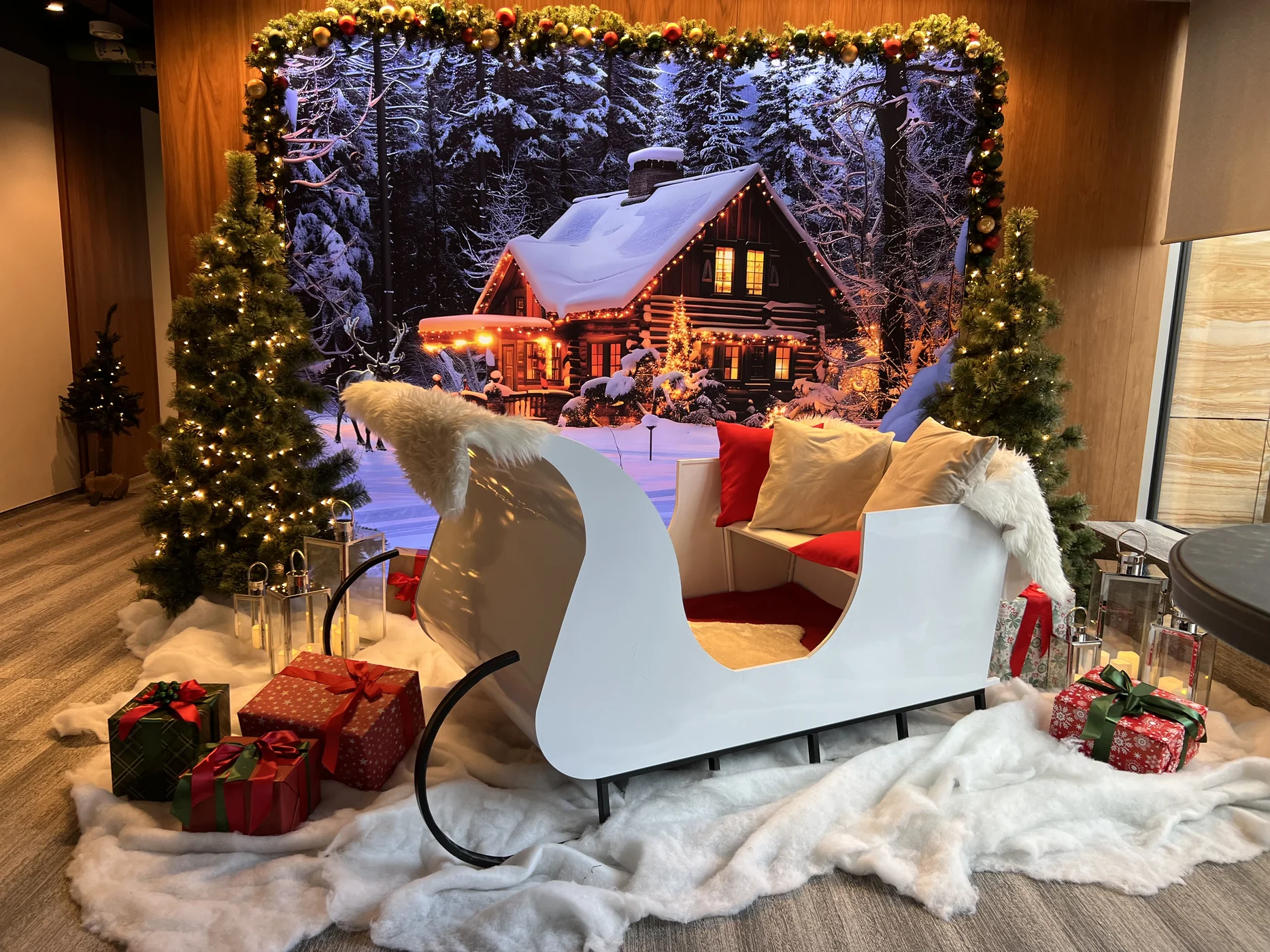 Kompleksowa zimowa scenografia z białymi saniami, czerwonymi i beżowymi poduszkami, futrzanymi narzutami oraz stylizowanymi prezentami ustawionymi na sztucznym śniegu, otoczona podświetlanymi choinkami i girlandą z bombkami, na tle wielkoformatowej grafiki przedstawiającej ośnieżony domek górski z bali w lesie – klimatyczne dekoracje idealne na firmowy event, galę świąteczną czy zimowy ślub. Profesjonalna wypożyczalnia z miasta Wrocław oferuje projekt, transport i montaż instalacji dostępnej w ofercie rent4events.pl oraz rent4weddings.pl, tworząc efektowną strefę foto i przestrzeń premium dopasowaną do charakteru wydarzenia - rent4events.pl wypożycz wrażenia, rent4wedding.pl zachowaj wspomnienia.