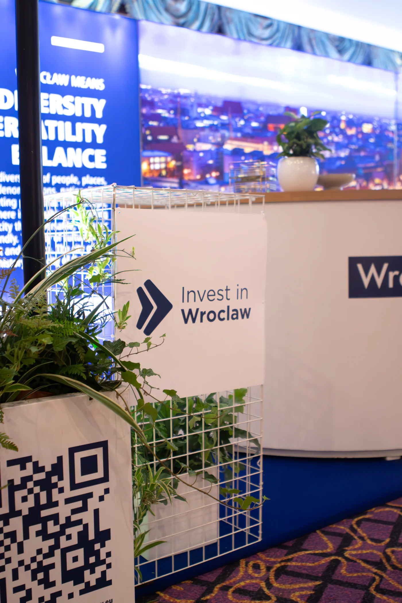 Stoisko targowe z brandingiem „Invest in Wrocław” przygotowane przez wypożyczalnia rent4events.pl i rent4weddings.pl to idealna scenografia na targi czy konferencje. Białe stelaże z dekoracjami roślinnymi, nowoczesne tablice, QR code oraz panorama miasta tworzą elegancką i funkcjonalną przestrzeń. Dekoracje i scenografia dostępne w wypożyczalni podnoszą prestiż każdej imprezy. Dla organizatorów eventów i uroczystości ślubnych we Wrocławiu rent4events.pl i rent4weddings.pl oferują kompleksowe rozwiązania - rent4events.pl wypożycz wrażenia, rent4weddings.pl zachowaj wspomnienia. Fot. @karolinakiec_fotografia.
