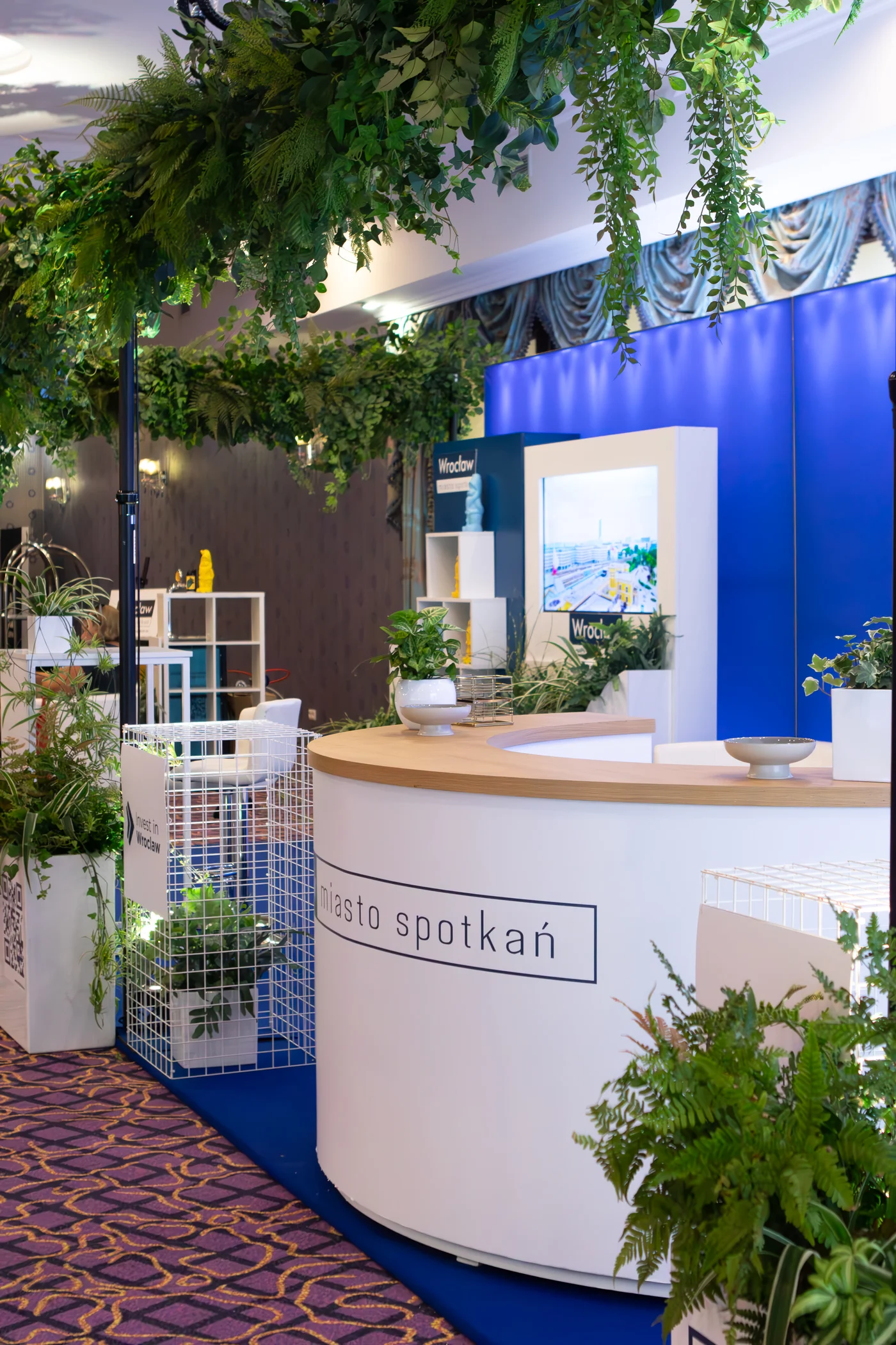 Stoisko eventowe „Wrocław – miasto spotkań” z dekoracjami od wypożyczalni rent4events.pl i rent4weddings.pl to nowoczesna scenografia na targi i konferencje. Elegancka lada, białe stelaże, bujne zielone dekoracje wiszące i stojące tworzą wyjątkową atmosferę. Połączenie roślin, nowoczesnych donic i ekranów multimedialnych sprawia, że każda przestrzeń eventowa staje się przyjazna i stylowa. Wypożyczalnia rent4events.pl oferuje dekoracje dopasowane do potrzeb zarówno eventowych, jak i ślubnych, gwarantując niezapomniane wrażenia - rent4events.pl wypożycz wrażenia, rent4weddings.pl zachowaj wspomnienia. Fot. @karolinakiec_fotografia.