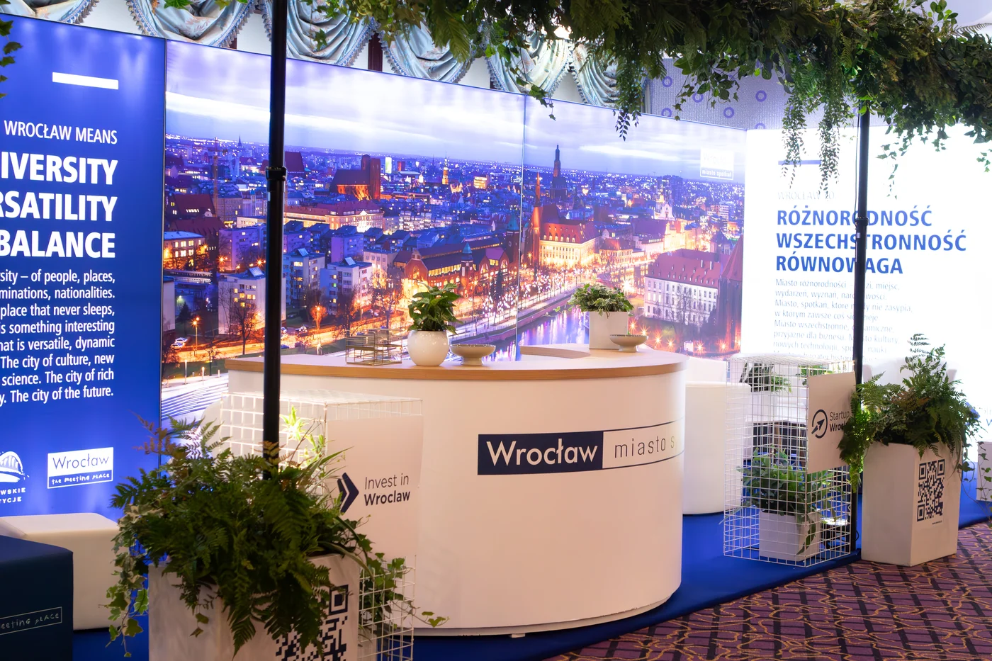 Stoisko eventowe „Wrocław – miasto spotkań” zrealizowane przez rent4events.pl / rent4weddings.pl. Elegancka lada, białe stelaże, zielone dekoracje roślinne oraz podświetlana panorama miasta tworzą niezwykłe wrażenie. Dekoracje, branding i elementy multimedialne przygotowane przez wypożyczalnię pozwalają zbudować przestrzeń, która podkreśla wartości miasta: różnorodność, wszechstronność i równowagę. Wypożyczalnia rent4events.pl oraz rent4weddings.pl – to gwarancja niezapomnianego wydarzenia - rent4events.pl wypożycz wrażenia, rent4weddings.pl zachowaj wspomnienia. Fot. @karolinakiec_fotografia.