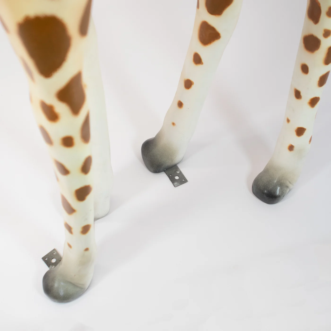 Duża, realistyczna figura żyrafy - zbliżenie na smukłe nogi w kolorze kremowym z brązowymi plamami oraz ciemnymi kopytami, z metalowymi elementami umożliwiającymi przykręcenie do podłoża - rent4events.pl wypożycz wrażenia, rent4wedding.pl zachowaj wspomnienia.