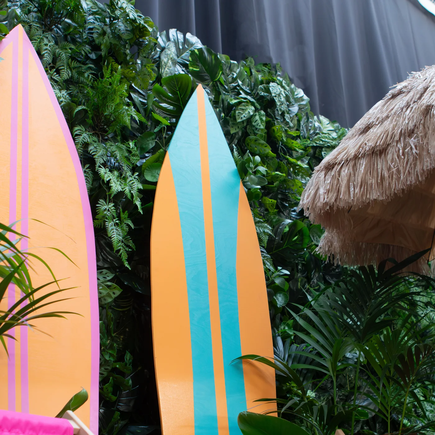 Tropikalna scenografia z pastelowymi deskami surfingowymi i jungle wall tworzy wyjątkowe dekoracje beach party dla nowoczesnych realizacji premium. Zbliżenie na fragment parasola typu tiki po prawej stranie oraz dwie kolorowe deski surfingowe, po lewej pomarańczowa z różową obramówką, w środku kadru deska w pomarańczowo-turkusowe pasy. Zielona ściana ze sztucznych liści tropikalnych, pastelowe kolory i naturalny parasol tiki podkreślają wakacyjny klimat oraz wysoką jakość aranżacji - rent4events.pl wypożycz wrażenia, rent4wedding.pl zachowaj wspomnienia.
